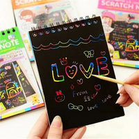 Black scratch art notebook with colorful scratch designs held by a hand, with other similar notebooks in the background.
