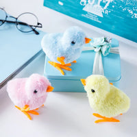 Three colorful plush chicks on a white surface with a blue box and glasses in the background.